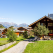 Traditional wooden mountain homes surrounded by lush green fields with scenic Himalayan peaks and a clear blue sky, representing peaceful countryside living in Himachal Pradesh.