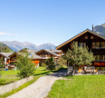 Traditional wooden mountain homes surrounded by lush green fields with scenic Himalayan peaks and a clear blue sky, representing peaceful countryside living in Himachal Pradesh.