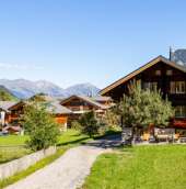 Traditional wooden mountain homes surrounded by lush green fields with scenic Himalayan peaks and a clear blue sky, representing peaceful countryside living in Himachal Pradesh.