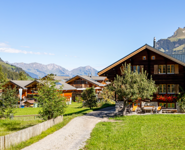 Traditional wooden mountain homes surrounded by lush green fields with scenic Himalayan peaks and a clear blue sky, representing peaceful countryside living in Himachal Pradesh.