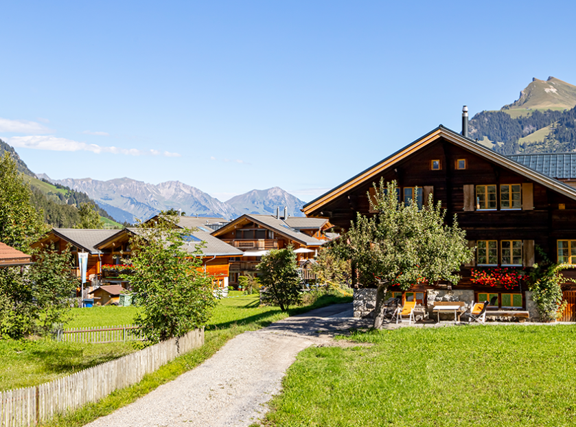 Traditional wooden mountain homes surrounded by lush green fields with scenic Himalayan peaks and a clear blue sky, representing peaceful countryside living in Himachal Pradesh.