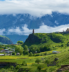 Scenic mountain valley landscape with lush green hills, a small village, a historic stone church, and mist-covered mountains in the background.