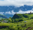 Scenic mountain valley landscape with lush green hills, a small village, a historic stone church, and mist-covered mountains in the background.