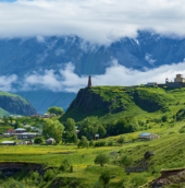 Scenic mountain valley landscape with lush green hills, a small village, a historic stone church, and mist-covered mountains in the background.
