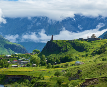 Scenic mountain valley landscape with lush green hills, a small village, a historic stone church, and mist-covered mountains in the background.