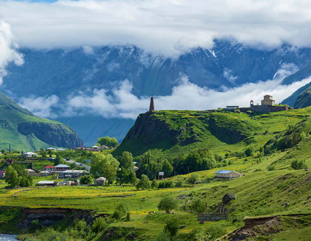 Scenic mountain valley landscape with lush green hills, a small village, a historic stone church, and mist-covered mountains in the background.