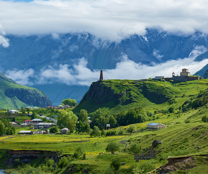 Scenic mountain valley landscape with lush green hills, a small village, a historic stone church, and mist-covered mountains in the background.