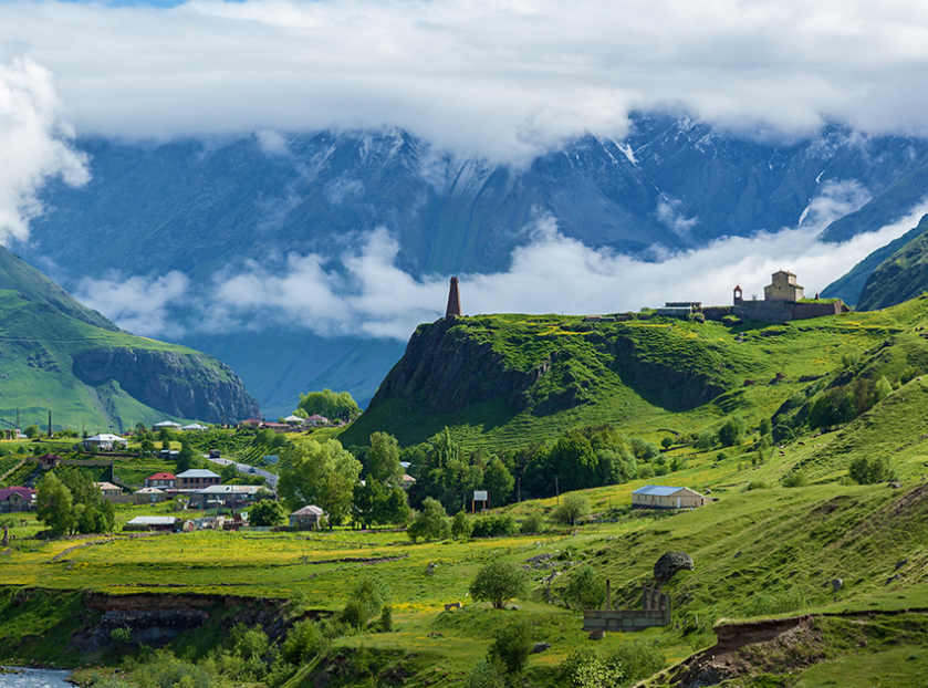Scenic mountain valley landscape with lush green hills, a small village, a historic stone church, and mist-covered mountains in the background.
