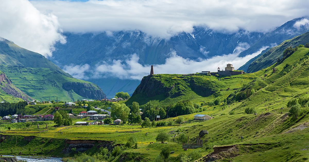 Scenic mountain valley landscape with lush green hills, a small village, a historic stone church, and mist-covered mountains in the background.