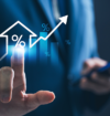 Business professional in a blue suit touching a glowing holographic interface showing a house icon with a percentage symbol, rising bar chart, and upward arrow, while holding a smartphone in the other hand.