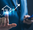Business professional in a blue suit touching a glowing holographic interface showing a house icon with a percentage symbol, rising bar chart, and upward arrow, while holding a smartphone in the other hand.