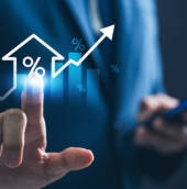 Business professional in a blue suit touching a glowing holographic interface showing a house icon with a percentage symbol, rising bar chart, and upward arrow, while holding a smartphone in the other hand.