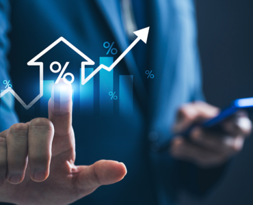 Business professional in a blue suit touching a glowing holographic interface showing a house icon with a percentage symbol, rising bar chart, and upward arrow, while holding a smartphone in the other hand.
