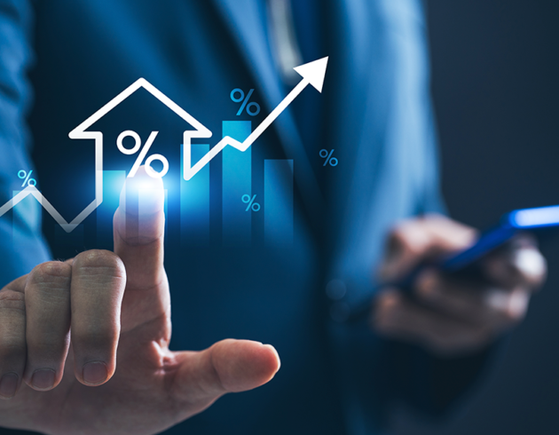 Business professional in a blue suit touching a glowing holographic interface showing a house icon with a percentage symbol, rising bar chart, and upward arrow, while holding a smartphone in the other hand.
