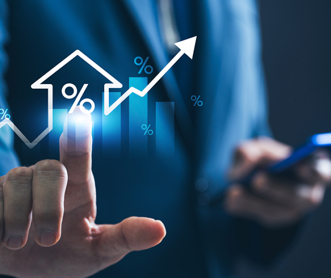Business professional in a blue suit touching a glowing holographic interface showing a house icon with a percentage symbol, rising bar chart, and upward arrow, while holding a smartphone in the other hand.