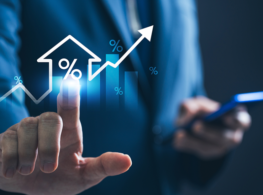 Business professional in a blue suit touching a glowing holographic interface showing a house icon with a percentage symbol, rising bar chart, and upward arrow, while holding a smartphone in the other hand.
