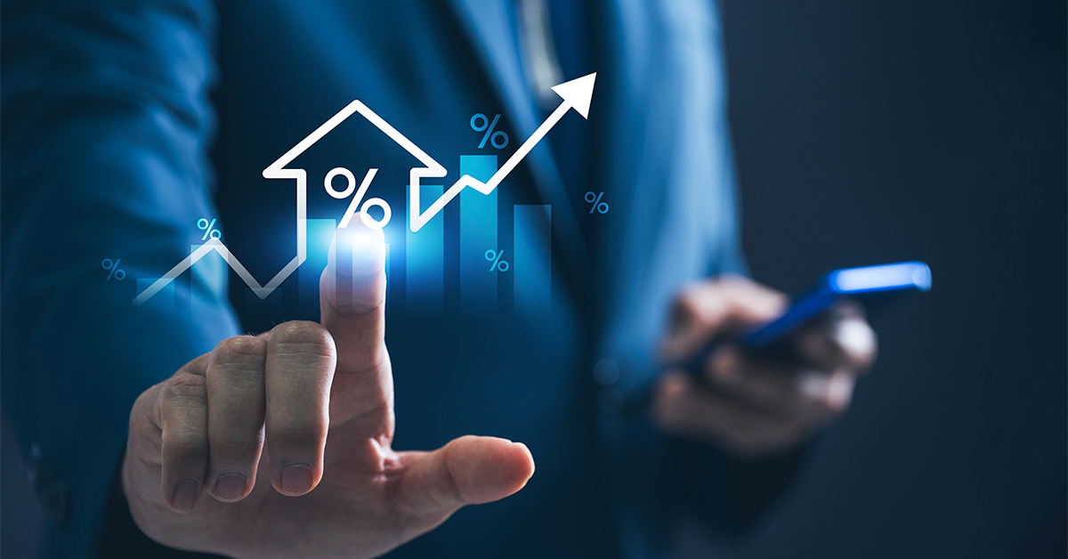 Business professional in a blue suit touching a glowing holographic interface showing a house icon with a percentage symbol, rising bar chart, and upward arrow, while holding a smartphone in the other hand.