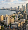 Mumbai city skyline from a high angle, with the Arabian Sea on the left, a curved coastline, and a dense mix of older buildings and tall skyscrapers filling the foreground and right side under a partly cloudy sky.