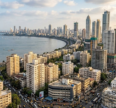 Mumbai city skyline from a high angle, with the Arabian Sea on the left, a curved coastline, and a dense mix of older buildings and tall skyscrapers filling the foreground and right side under a partly cloudy sky.
