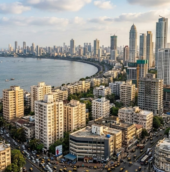 Mumbai city skyline from a high angle, with the Arabian Sea on the left, a curved coastline, and a dense mix of older buildings and tall skyscrapers filling the foreground and right side under a partly cloudy sky.
