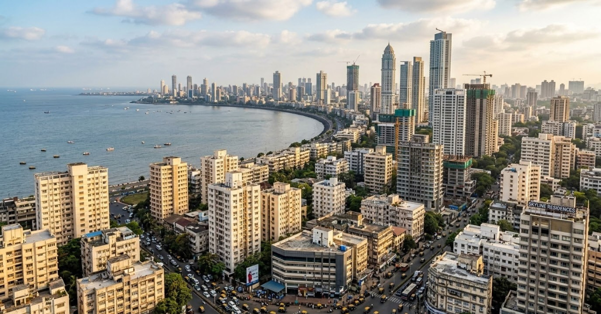 Mumbai city skyline from a high angle, with the Arabian Sea on the left, a curved coastline, and a dense mix of older buildings and tall skyscrapers filling the foreground and right side under a partly cloudy sky.