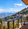 A luxurious hotel balcony with two woven armchairs and a small round table overlooking a hillside town of colorful buildings and green trees, with a yellow church tower on the left, dense pine forests, and a vast range of snow-capped mountains under a bright blue sky.