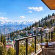 A luxurious hotel balcony with two woven armchairs and a small round table overlooking a hillside town of colorful buildings and green trees, with a yellow church tower on the left, dense pine forests, and a vast range of snow-capped mountains under a bright blue sky.