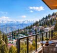 A luxurious hotel balcony with two woven armchairs and a small round table overlooking a hillside town of colorful buildings and green trees, with a yellow church tower on the left, dense pine forests, and a vast range of snow-capped mountains under a bright blue sky.