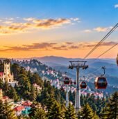 Shimla ropeway with red cable cars overlooking the city, pine forests, and Himalayan mountains at sunset.