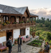 Stone-built hilltop homestay in Uttarakhand featuring a traditional wooden balcony, colorful flowers, and views of green hills and snow-capped Himalayan mountains.