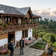 Stone-built hilltop homestay in Uttarakhand featuring a traditional wooden balcony, colorful flowers, and views of green hills and snow-capped Himalayan mountains.