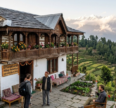 Stone-built hilltop homestay in Uttarakhand featuring a traditional wooden balcony, colorful flowers, and views of green hills and snow-capped Himalayan mountains.