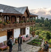Stone-built hilltop homestay in Uttarakhand featuring a traditional wooden balcony, colorful flowers, and views of green hills and snow-capped Himalayan mountains.