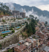 Luxury villas and apartments in Kasauli overlooking a misty mountain landscape, contrasted with densely packed residential buildings and ongoing real estate development in the hills.
