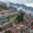 Luxury villas and apartments in Kasauli overlooking a misty mountain landscape, contrasted with densely packed residential buildings and ongoing real estate development in the hills.