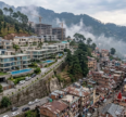Luxury villas and apartments in Kasauli overlooking a misty mountain landscape, contrasted with densely packed residential buildings and ongoing real estate development in the hills.