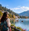 A solo female traveler with a backpack enjoys a scenic lakeside view, surrounded by mountains, colorful hillside buildings, and boats on the water.