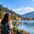 A solo female traveler with a backpack enjoys a scenic lakeside view, surrounded by mountains, colorful hillside buildings, and boats on the water.