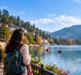 A solo female traveler with a backpack enjoys a scenic lakeside view, surrounded by mountains, colorful hillside buildings, and boats on the water.