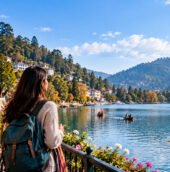A solo female traveler with a backpack enjoys a scenic lakeside view, surrounded by mountains, colorful hillside buildings, and boats on the water.