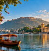 Scenic golden-hour view of a calm lake in Rajasthan, India, with a traditional wooden boat under a red canopy in the foreground. On the left, a stone walkway lined with pink bougainvillea flowers and an ornate building; on the right, a grand multi-domed palace complex reflected in the water; in the background, a large hilltop fort under a bright blue sky with soft clouds.
