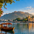 Scenic golden-hour view of a calm lake in Rajasthan, India, with a traditional wooden boat under a red canopy in the foreground. On the left, a stone walkway lined with pink bougainvillea flowers and an ornate building; on the right, a grand multi-domed palace complex reflected in the water; in the background, a large hilltop fort under a bright blue sky with soft clouds.
