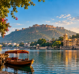 Scenic golden-hour view of a calm lake in Rajasthan, India, with a traditional wooden boat under a red canopy in the foreground. On the left, a stone walkway lined with pink bougainvillea flowers and an ornate building; on the right, a grand multi-domed palace complex reflected in the water; in the background, a large hilltop fort under a bright blue sky with soft clouds.