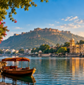 Scenic golden-hour view of a calm lake in Rajasthan, India, with a traditional wooden boat under a red canopy in the foreground. On the left, a stone walkway lined with pink bougainvillea flowers and an ornate building; on the right, a grand multi-domed palace complex reflected in the water; in the background, a large hilltop fort under a bright blue sky with soft clouds.