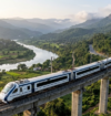 Vande Bharat train traveling on an elevated railway bridge through lush green mountains, overlooking a winding river and scenic valley.